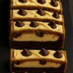 A stack of Brown Butter Chocolate Chip Cookie Cheesecake Bars on a marble table, showing creamy cheesecake and golden cookie layers.