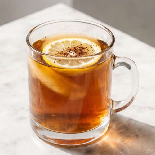 Warm natural morning drink in a clear glass mug with lemon slices and cinnamon on top, sitting on a bright kitchen counter.