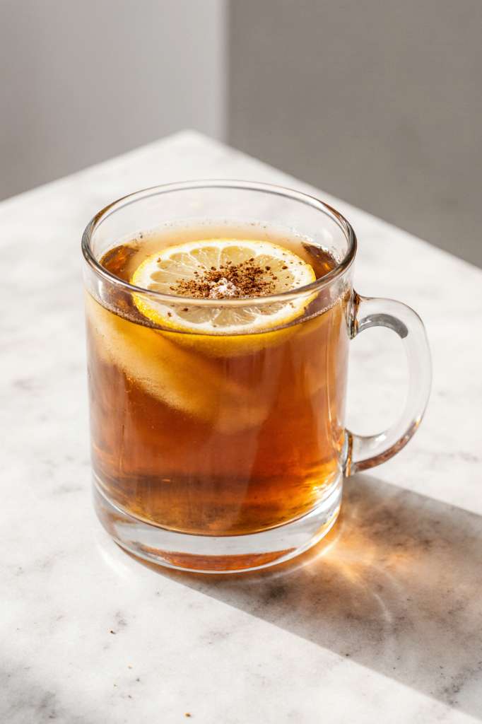 Warm natural morning drink in a clear glass mug with lemon slices and cinnamon on top, sitting on a bright kitchen counter.